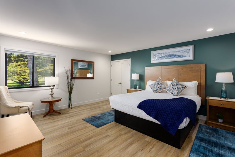 Modern bedroom with a large bed, blue accents, two bedside tables with lamps, a chair near a window, a mirror, and wooden flooring—reminiscent of the elegant yet relaxed style found in many Moonstone Beach Cambria CA hotel rooms. A landscape picture hangs above the bed, adding to the tranquil coastal vibe.