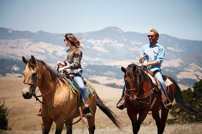 Two people riding horses on a sunny day in a grassy field with mountains in the background, reminiscent of the serene landscapes near Moonstone Beach Cambria CA hotel. Both riders are casually dressed and appear to be enjoying the outdoor scenery.